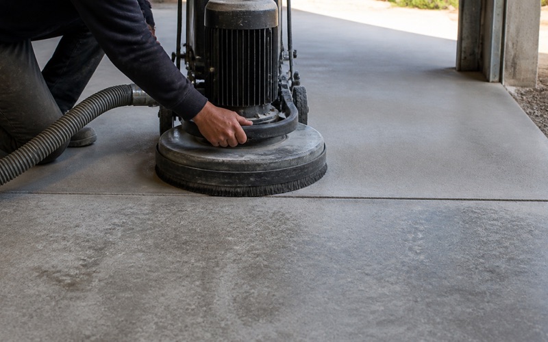 An applicator smooths gray coating up the edge of a concrete equipment pad in a service corridor beside an open trench drain.
