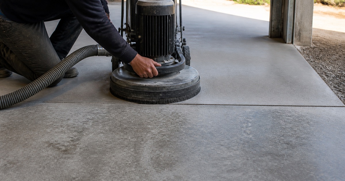 An applicator smooths gray coating up the edge of a concrete equipment pad in a service corridor beside an open trench drain.