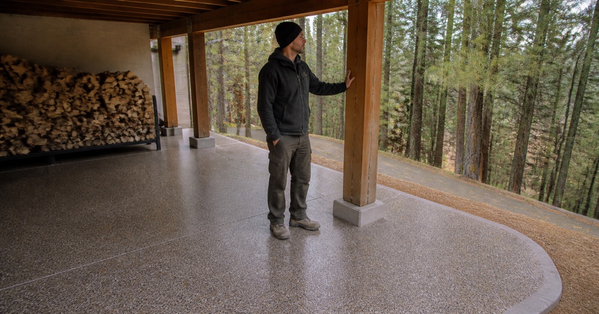 A worker in mittens rests a hand flat on the cold bare slab of a snowy mountain cabin garage, where a finished flake corner meets the wood door threshold.