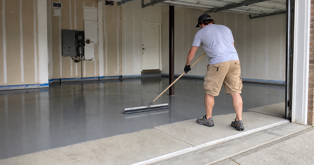 A kneeling applicator presses urethane cement into a coved base along a commercial kitchen wall with a floor drain nearby.